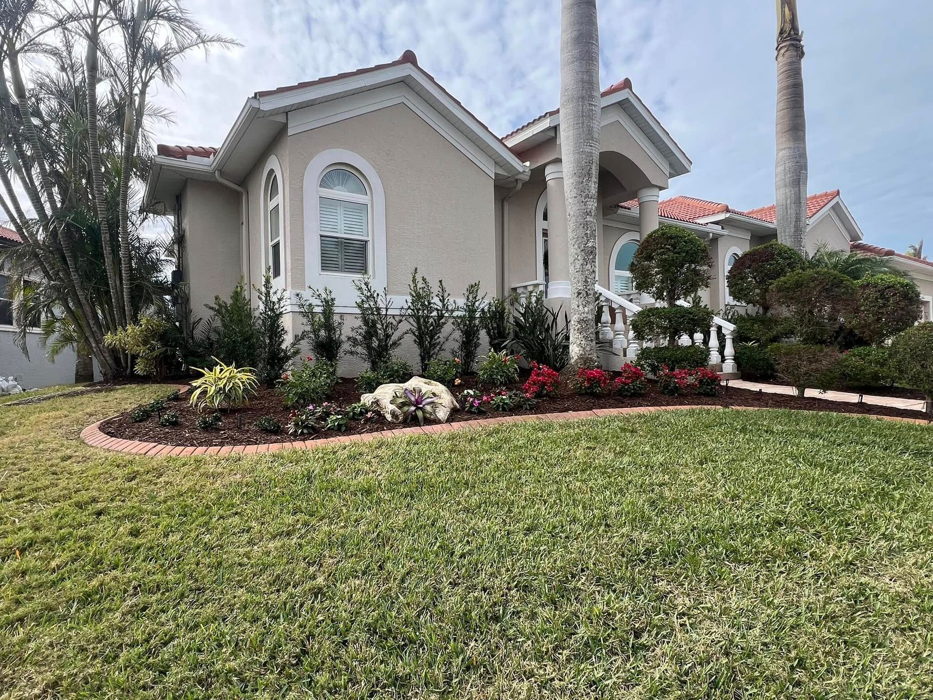 Beige house with red tile roof, lush landscaping in front.