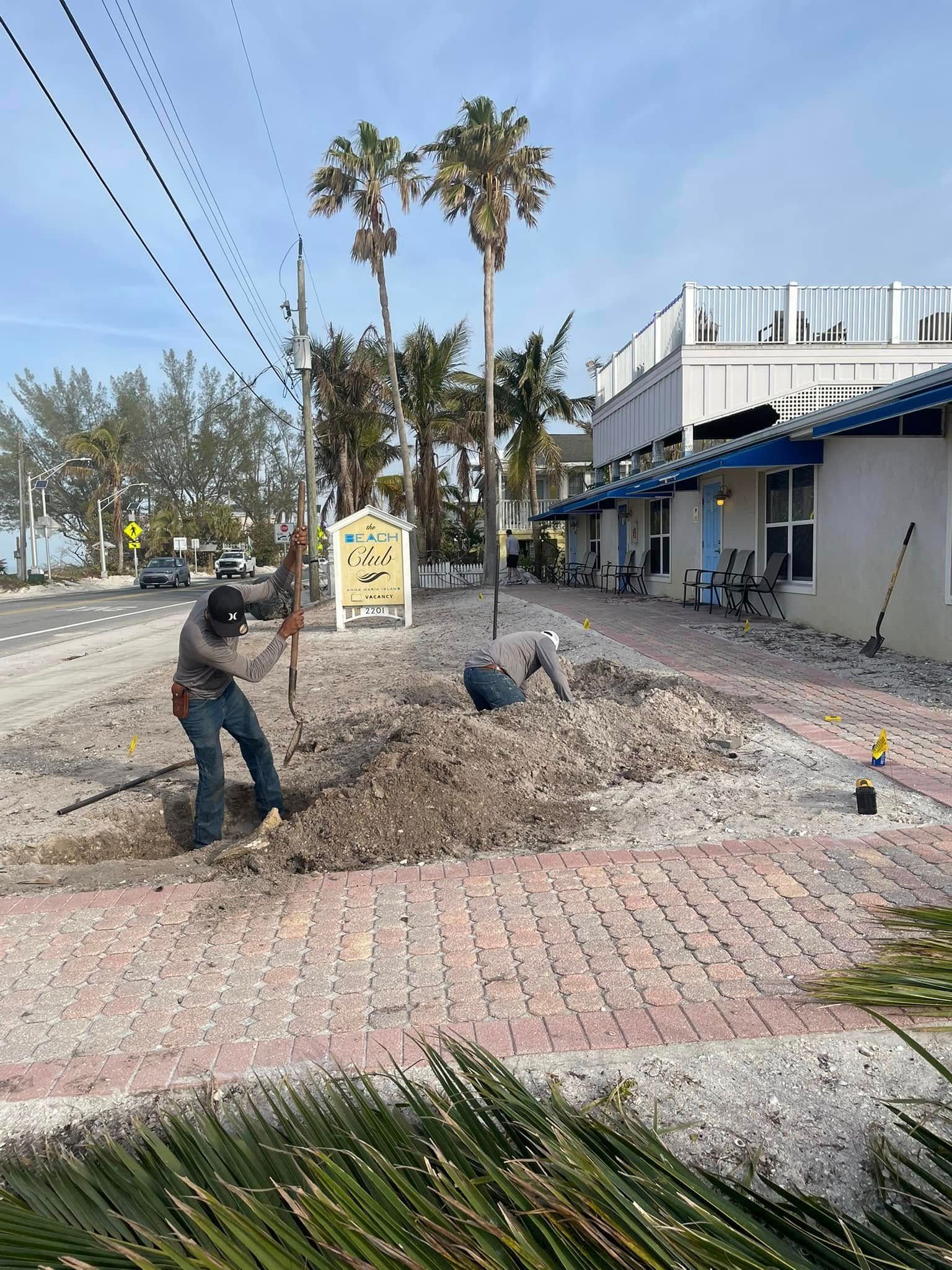 Two workers digging in a gravel area on a brick sidewalk near a beach and building.