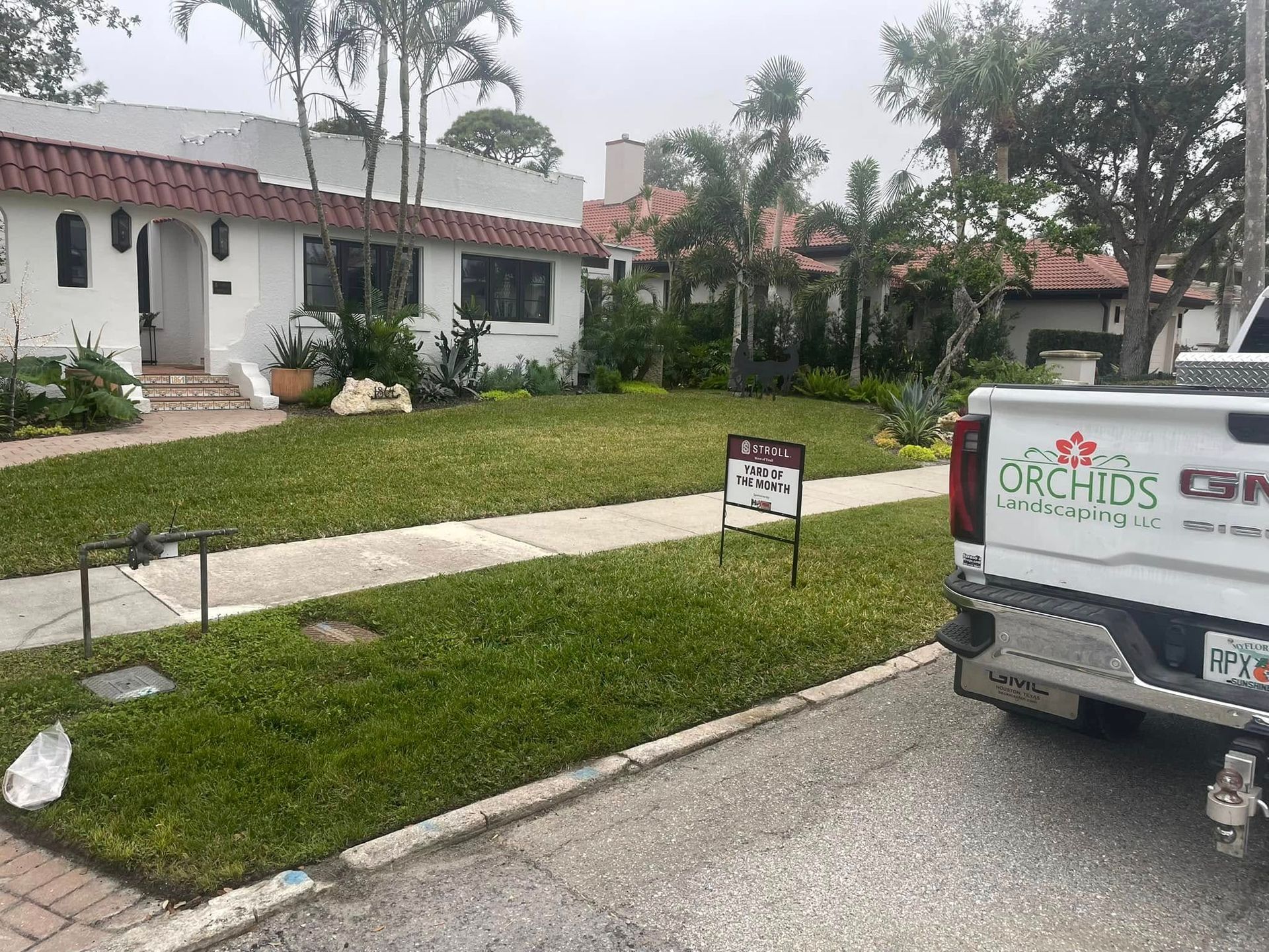 White house with red tile roof, sign in lawn, and a parked GMC truck from Orchids in front.