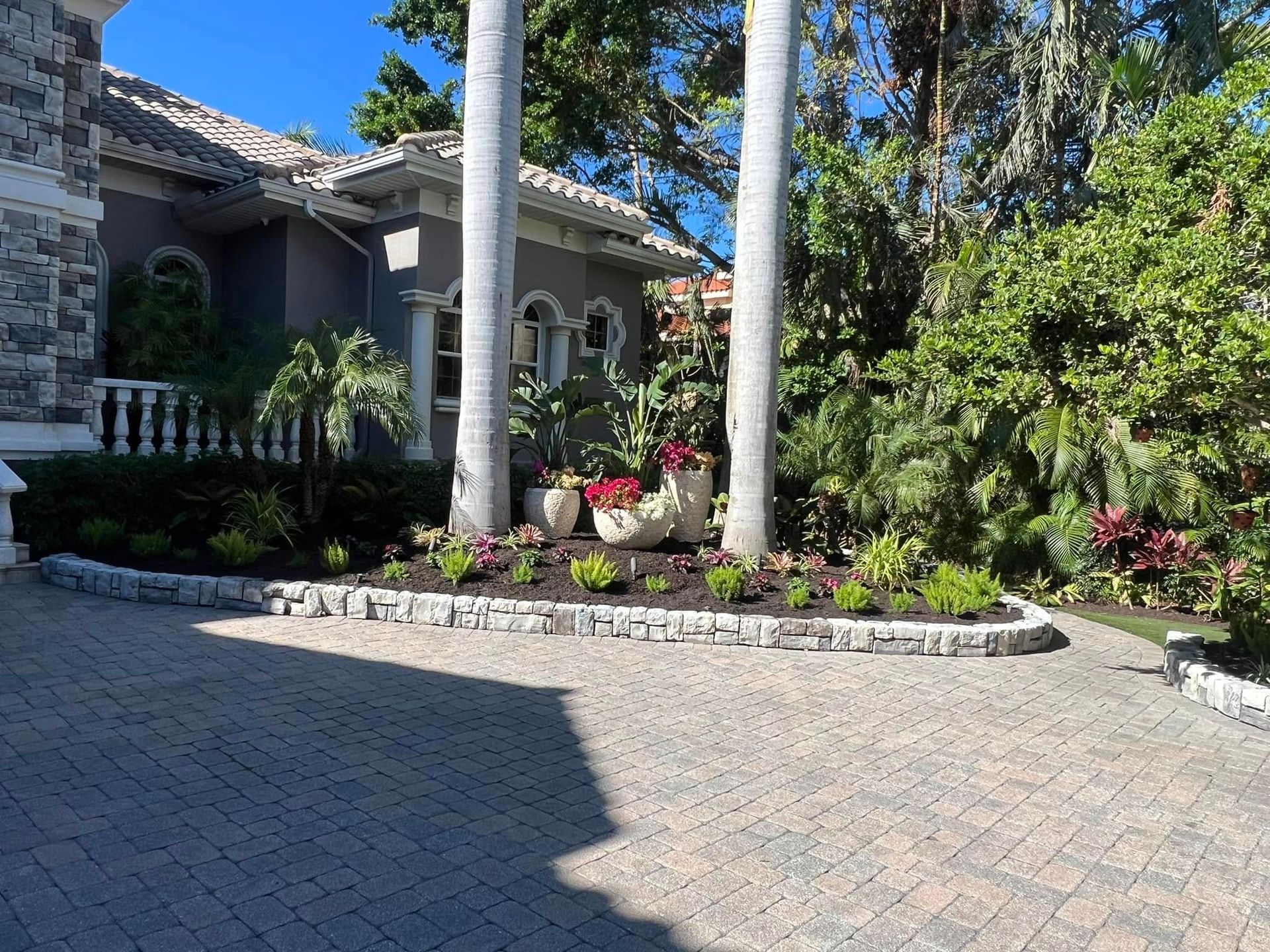 Driveway with manicured landscaping, palm trees, and a gray house.