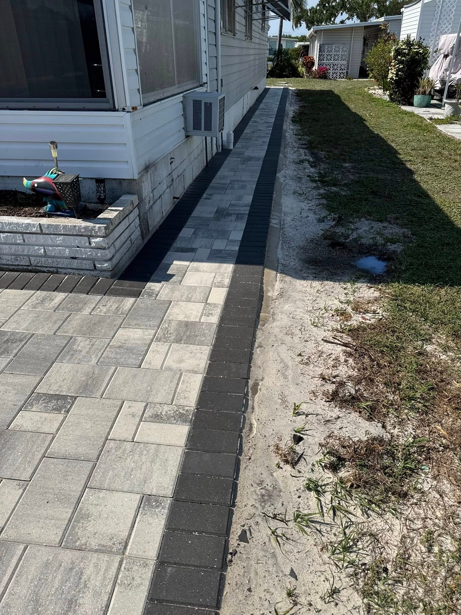 Brick walkway with dark border alongside a white house, transitioning to a sandy area and grass.