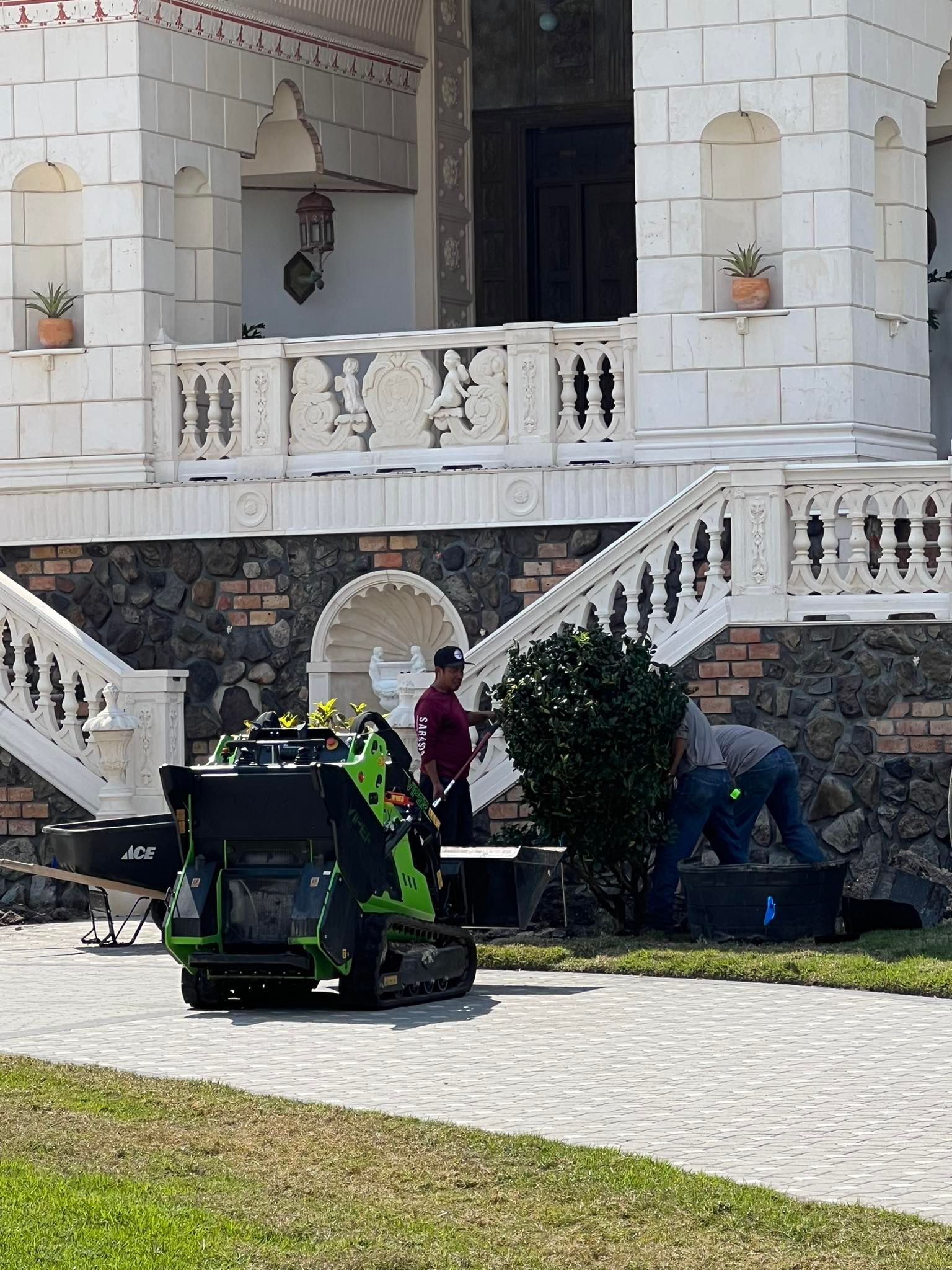 A mini excavator next to a building with people working near steps and a hedge.