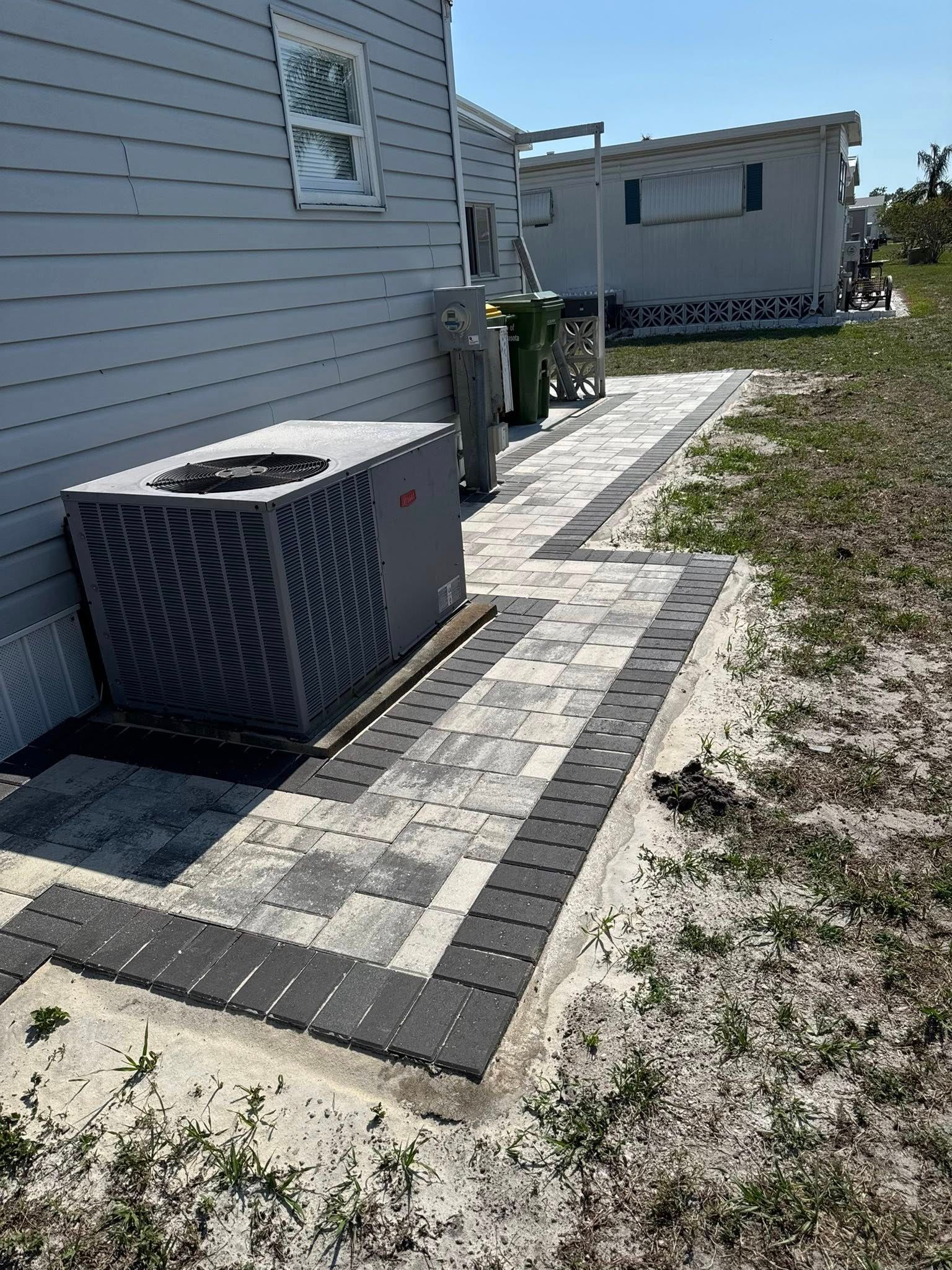 A paved walkway with a brick border next to a gray house and an AC unit.