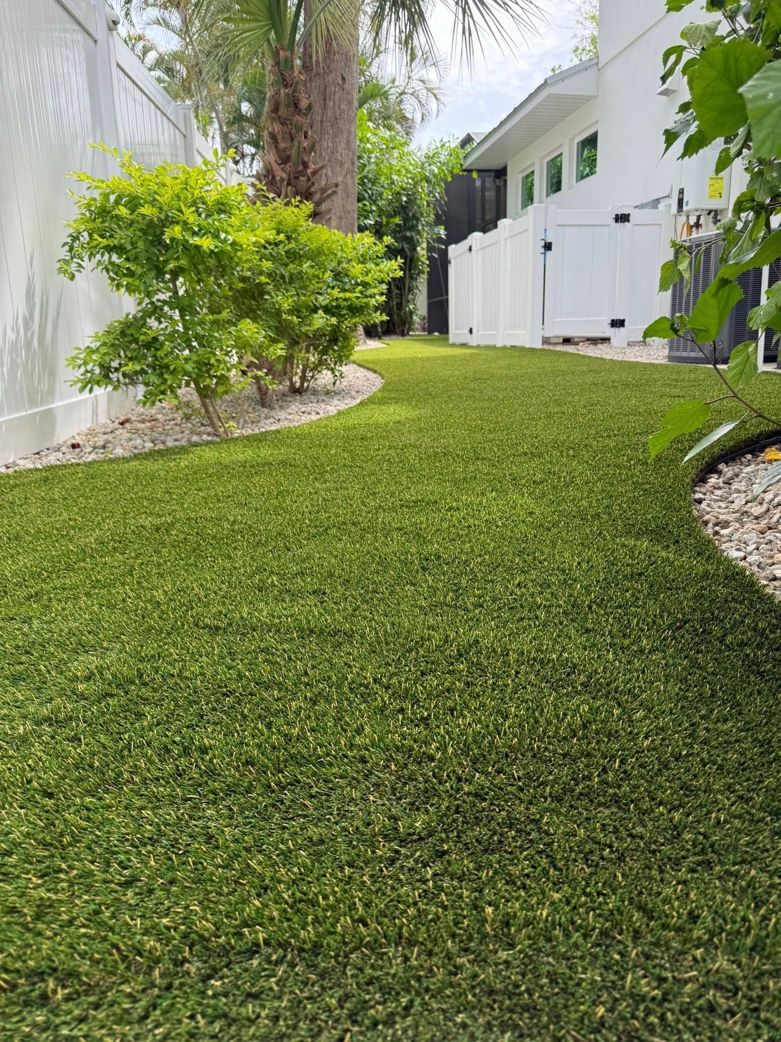 Green artificial turf in a backyard, edged by white fencing and a green bush.