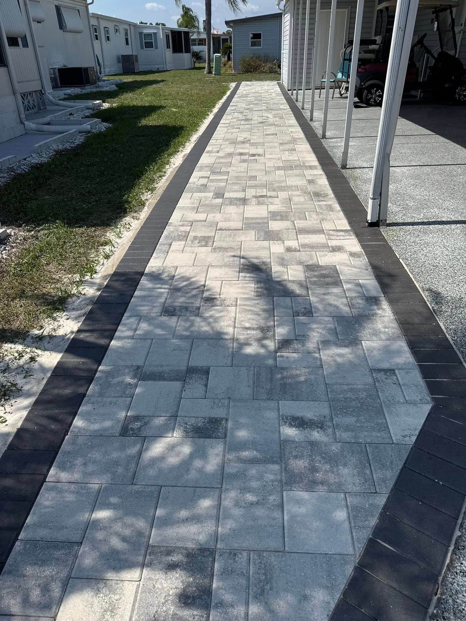 Paved walkway with gray and black pavers, flanked by grass and a building with a white awning.