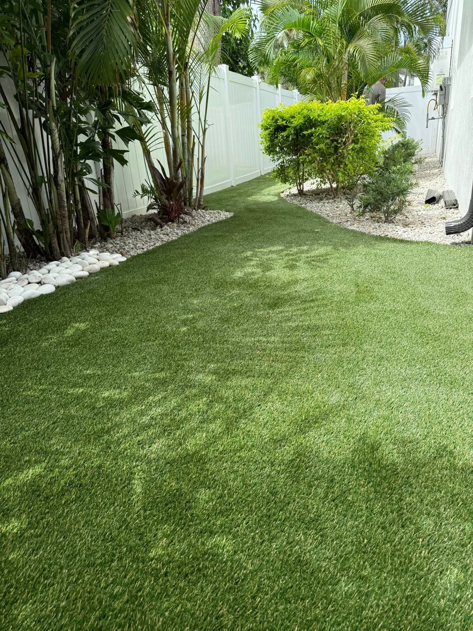Green artificial turf in a backyard, bordered by a white fence, bushes, and white stones.