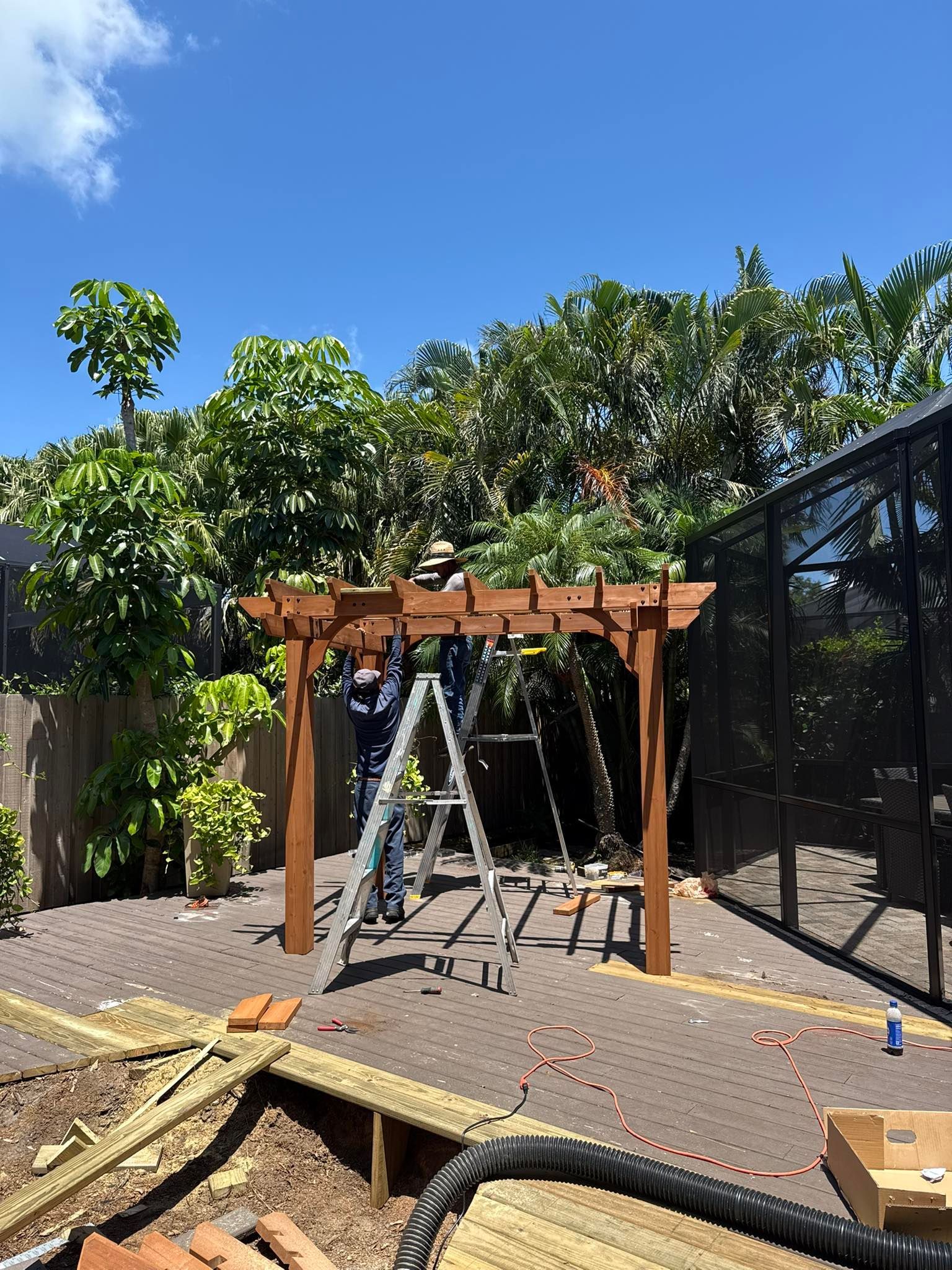 Workers building a brown wooden pergola in a backyard, using ladders. Sunny day, greenery in the background.