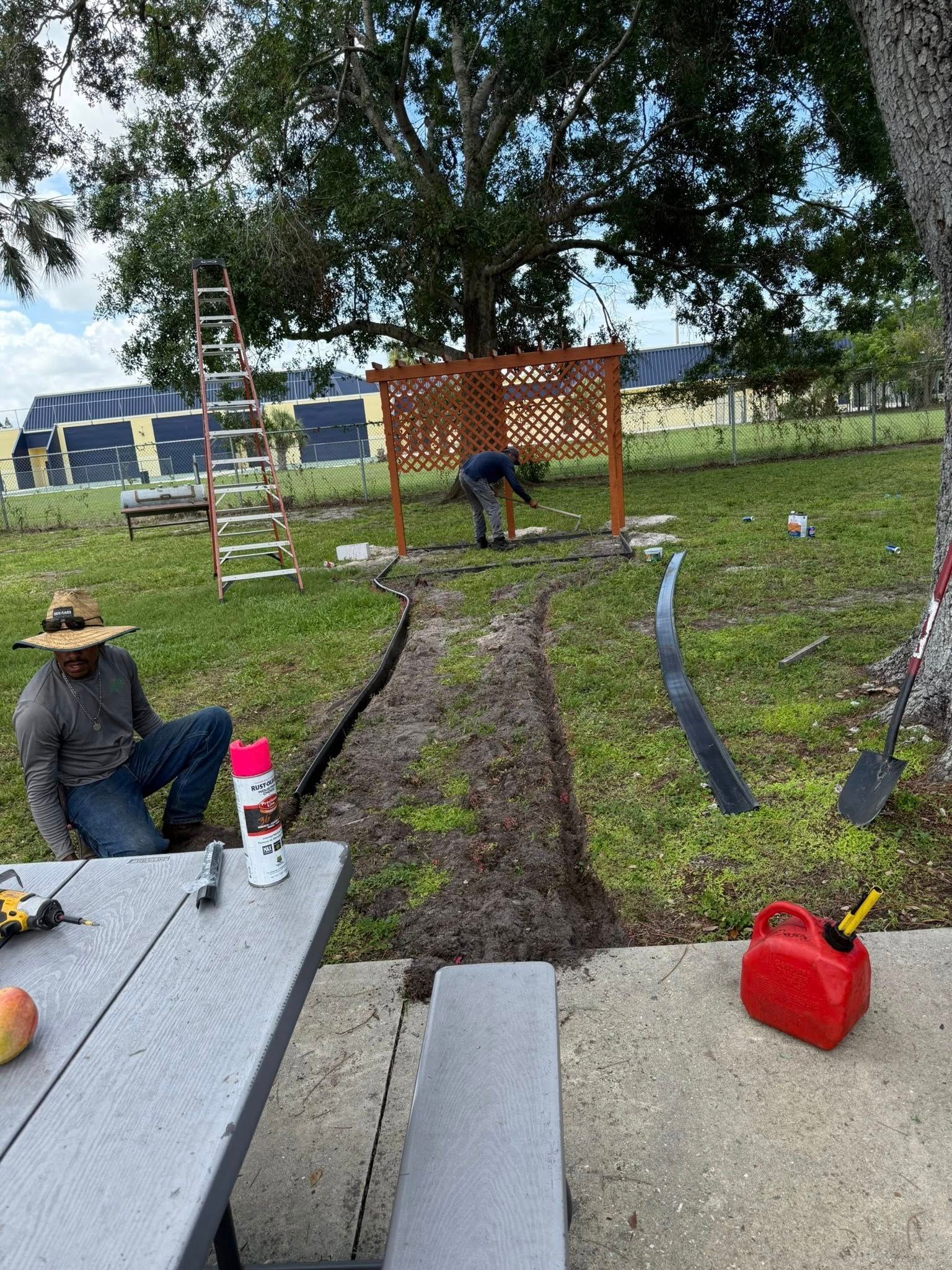 Workers installing a pathway with a decorative fence in a grassy yard, with tools and a picnic table present.