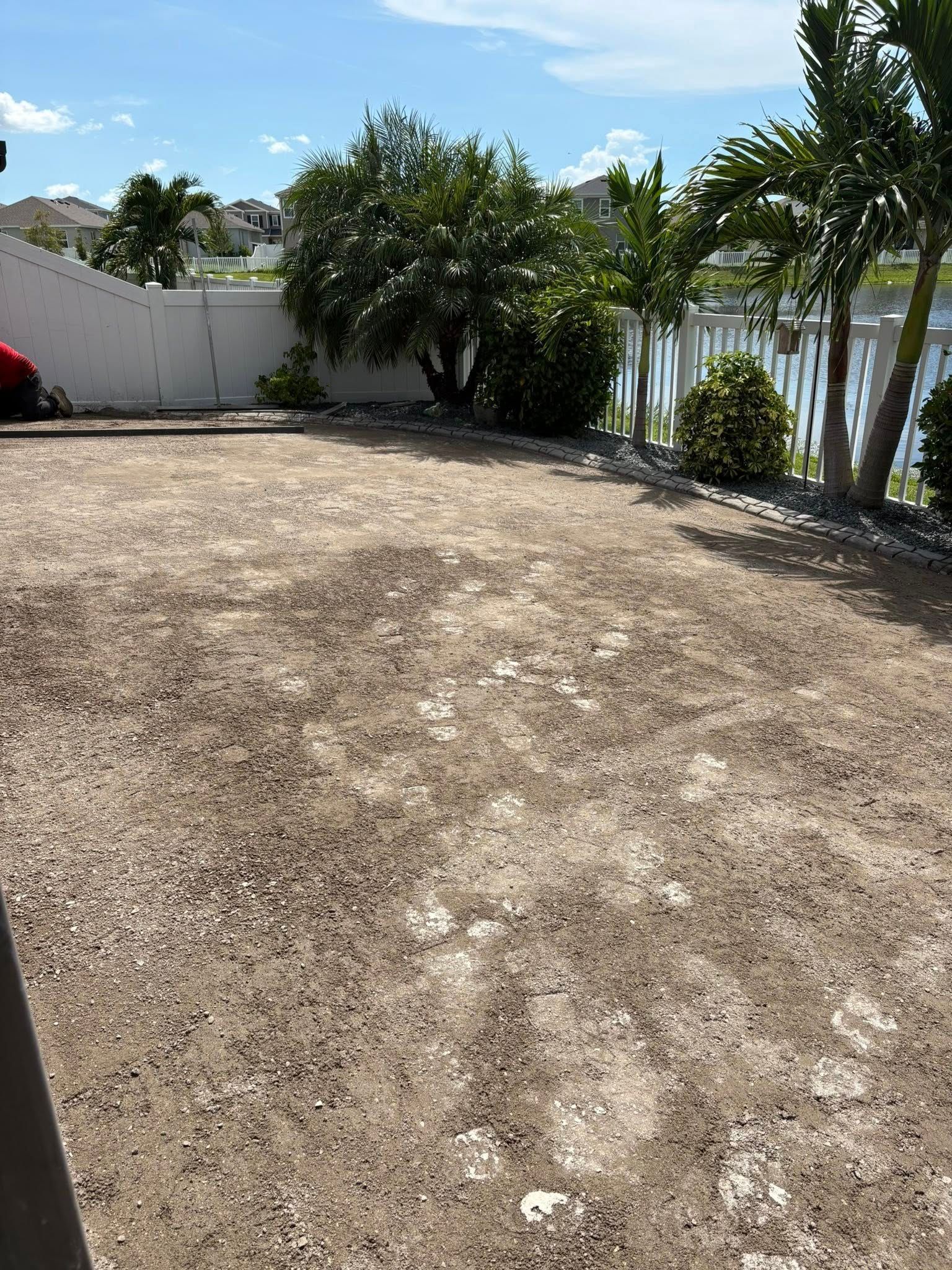 A gravel yard with a white fence, bushes, and palm trees, overlooking a body of water on a sunny day.