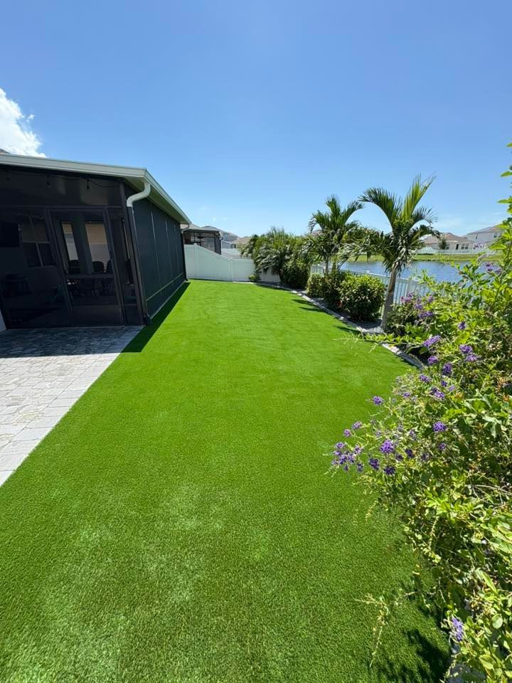 Lush green artificial turf in backyard, next to patio, white fence, and lake. Clear, sunny sky.