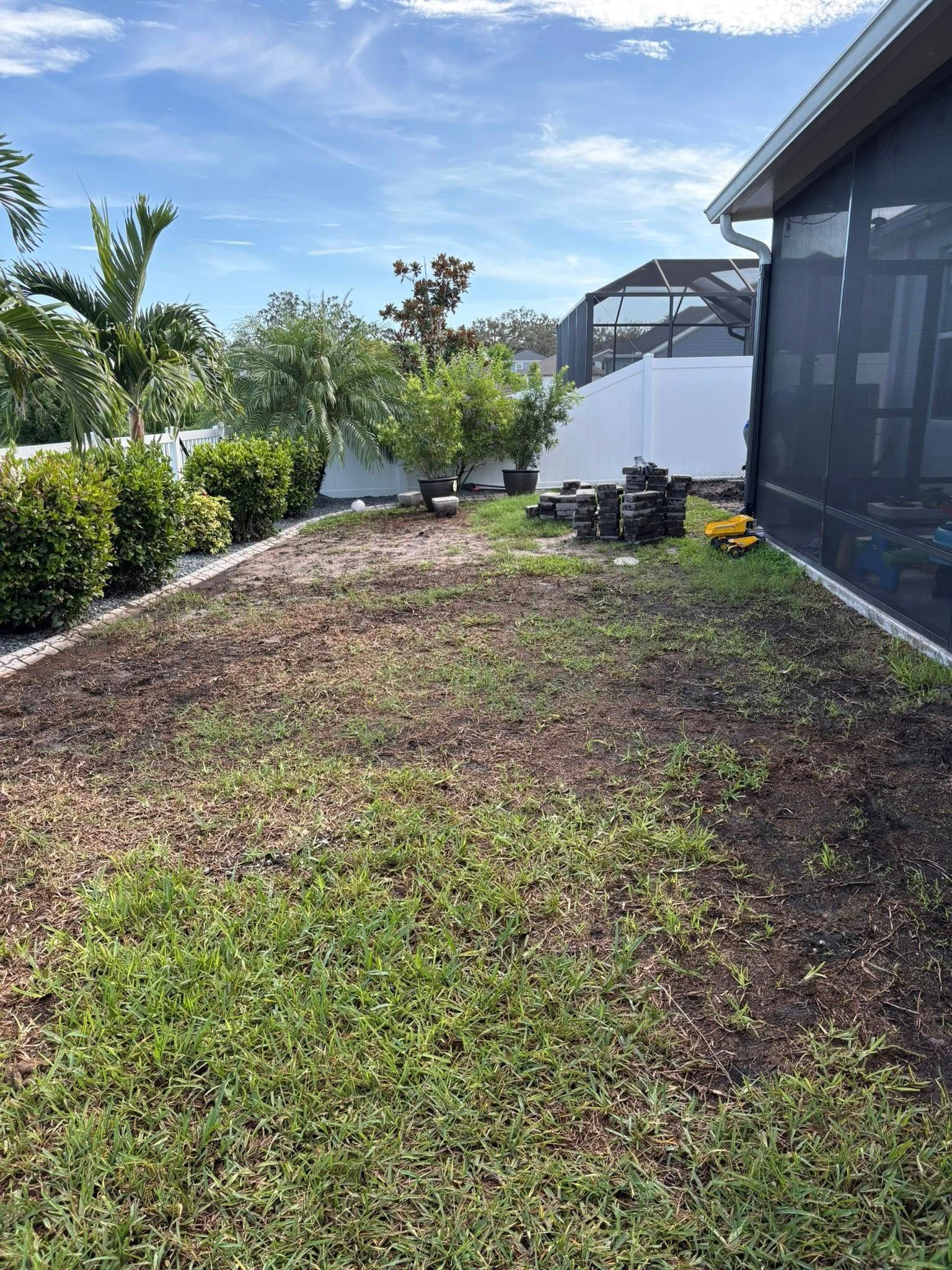 Backyard with lawn, garden beds, white fence, and house with screen enclosure under a blue sky.
