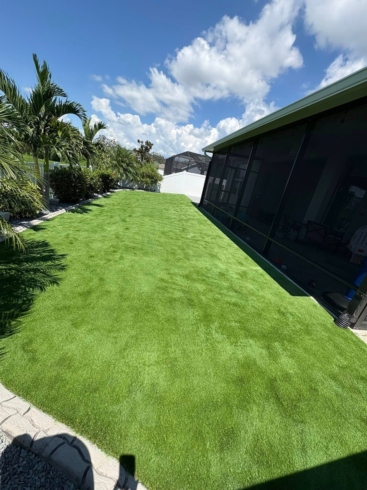 Green artificial turf sloping down to a water view, adjacent to a screened-in enclosure. Blue sky with clouds.