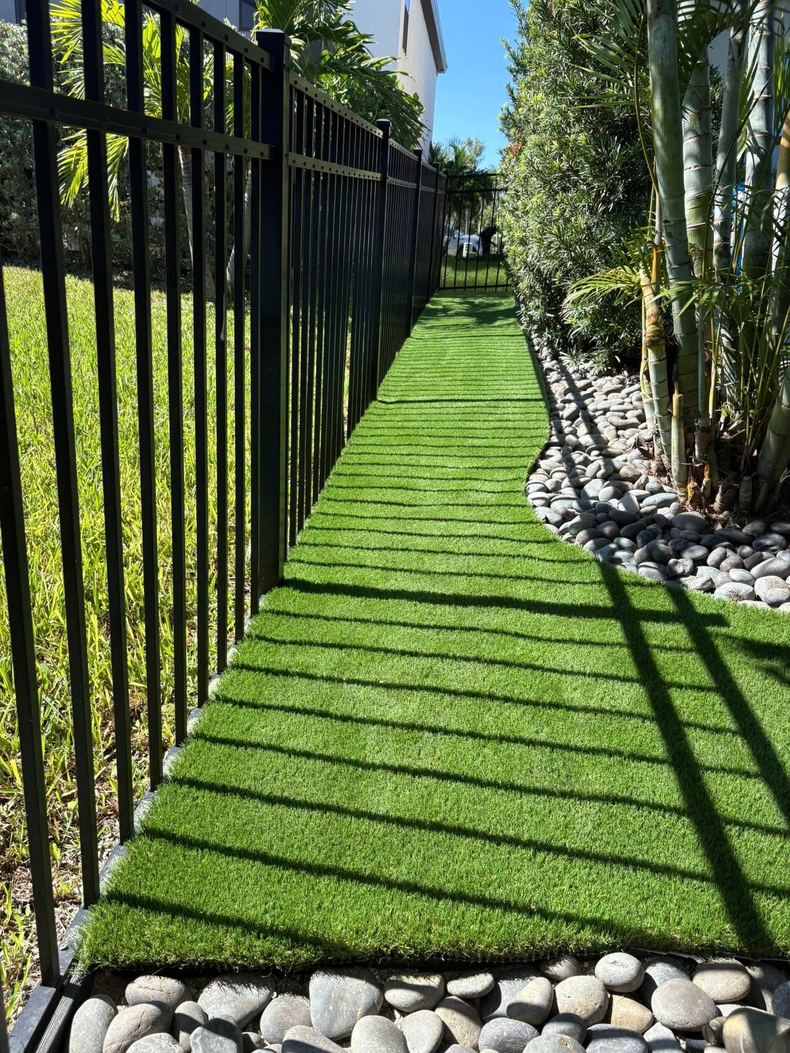 A narrow strip of artificial turf pathway bordered by a black fence and pebbles, beside trees.