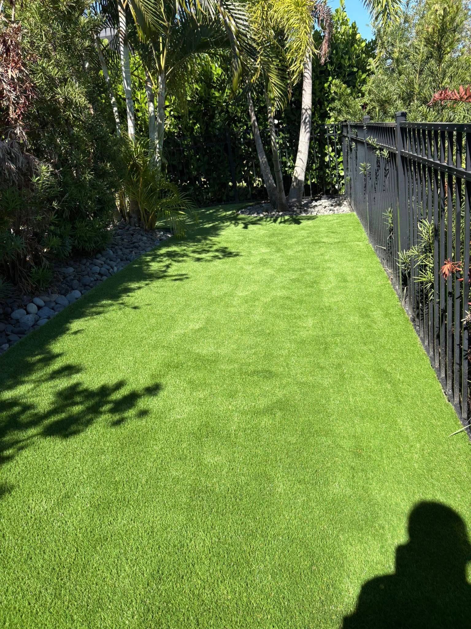 Green artificial grass next to a black fence, with trees and shadows.