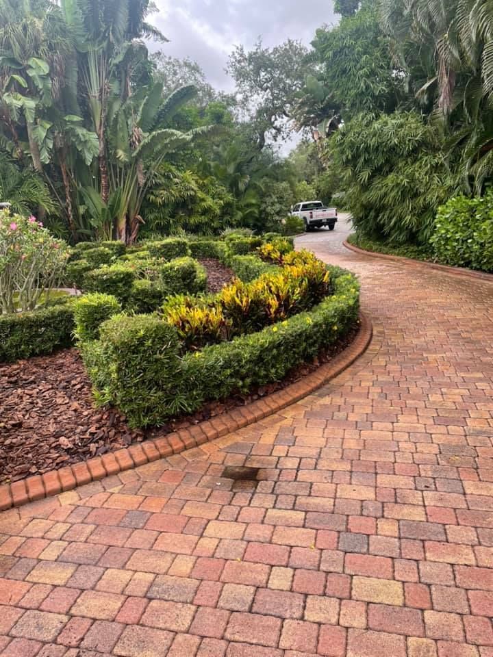 Brick driveway curves through lush, landscaped garden with trimmed hedges and colorful foliage.