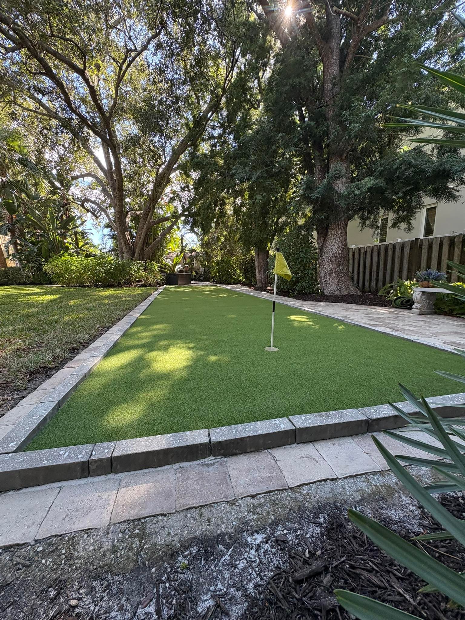 Artificial turf putting green in a backyard, edged with stone pavers and bordering landscaping, with a flag and trees.