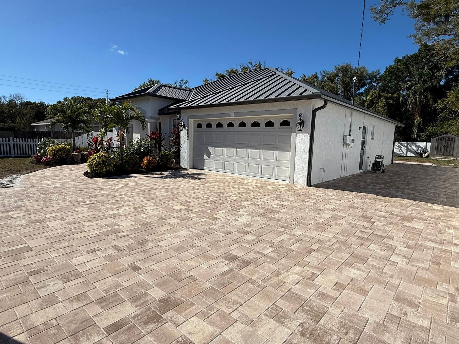 White house with brick driveway, garage, and landscaping under a blue sky.