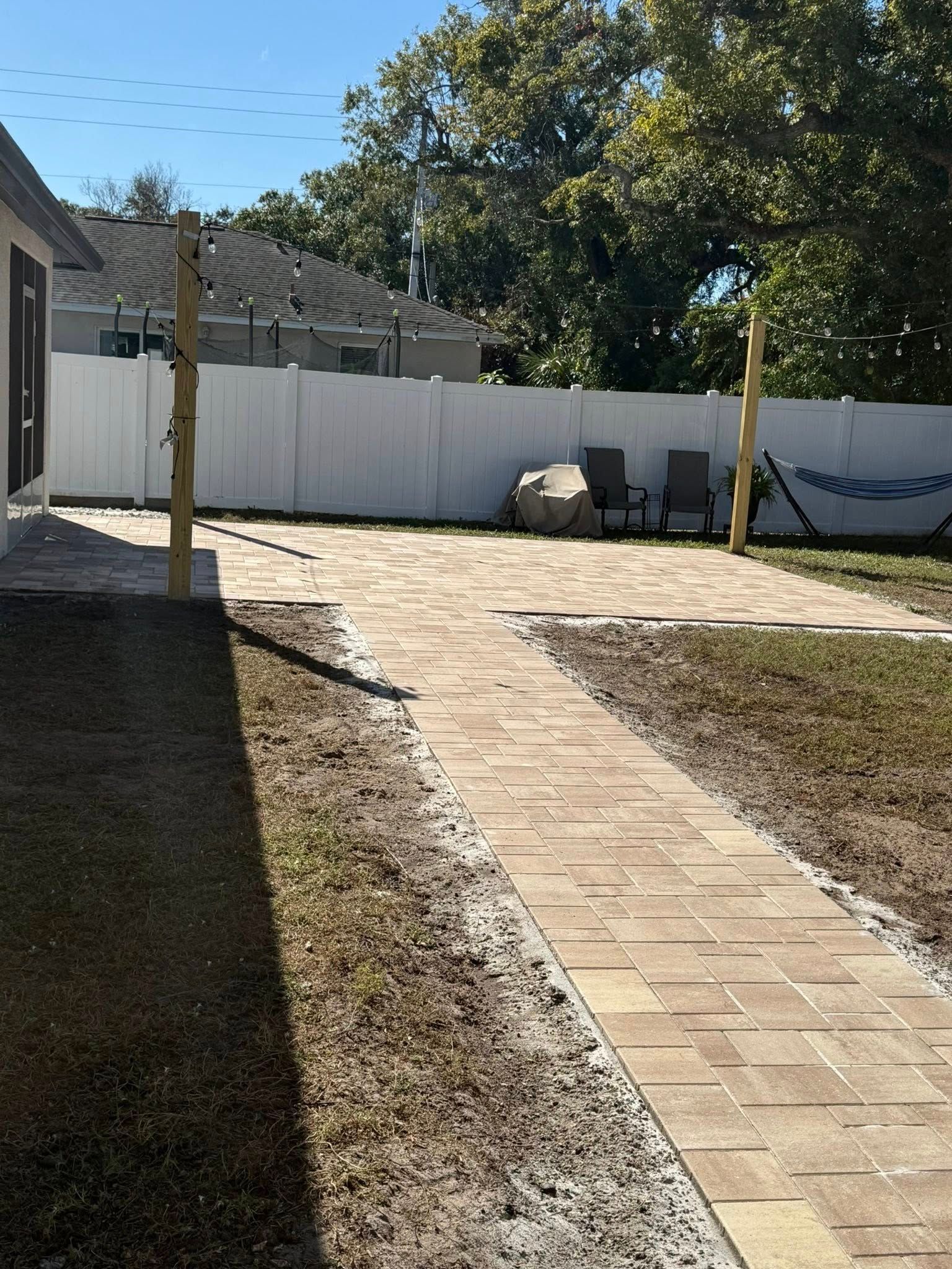Pathway of pavers through a backyard to a patio area with a white fence.