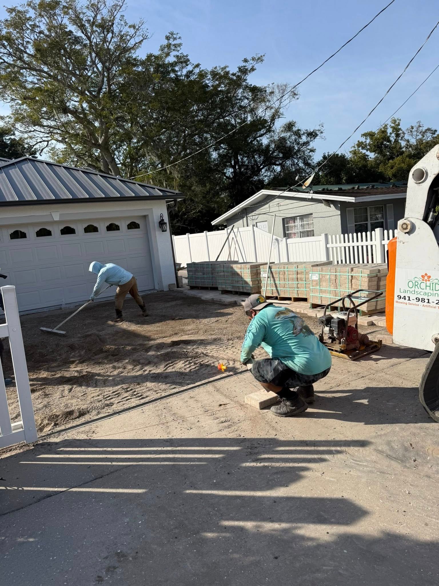 Two workers prepping a yard with rakes and tools near a garage and stacked bricks. Sunny day.