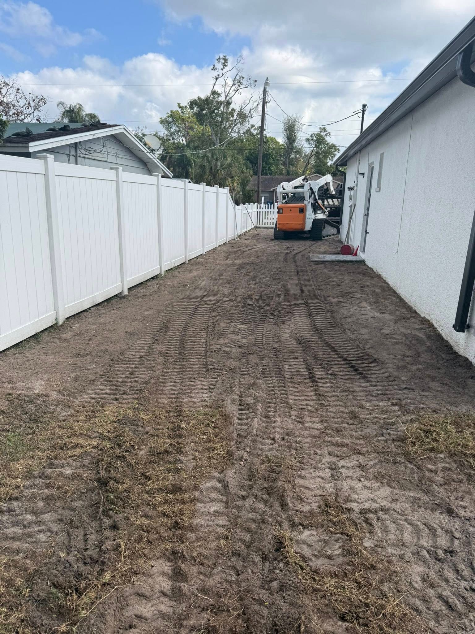 A dirt patch beside a white fence and a building. A small bobcat sits in the distance.