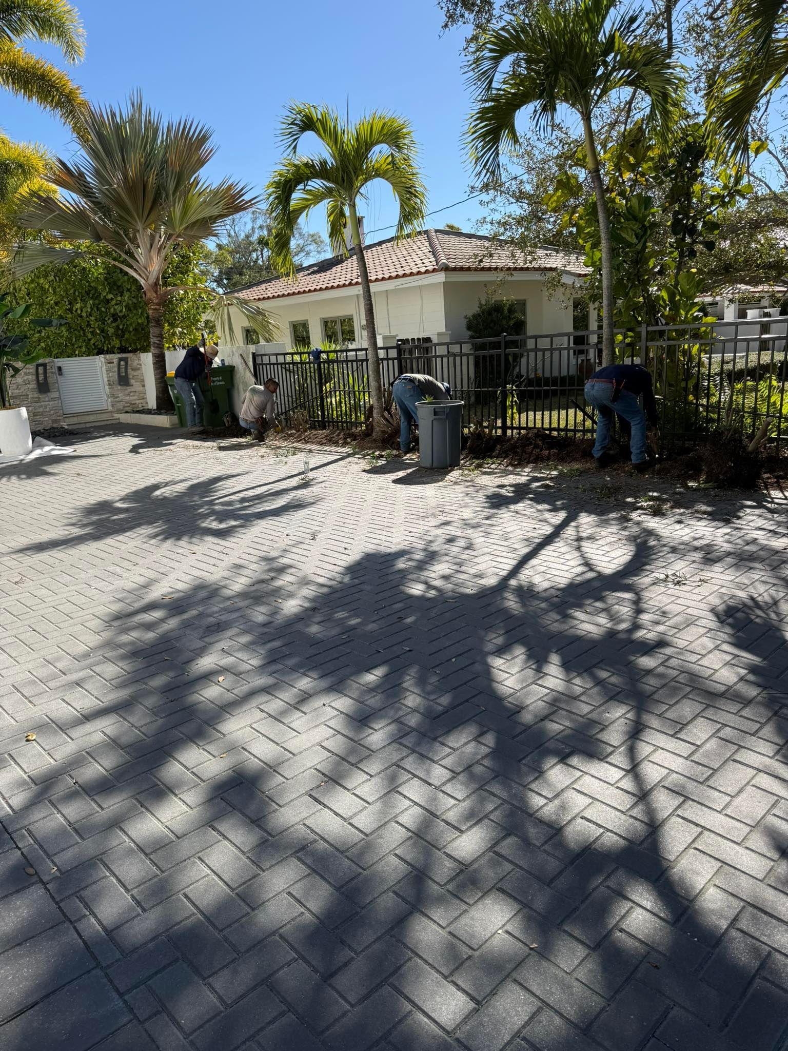 Workers cleaning a paved outdoor area near a small building with a tiled roof, trees, and metal fencing.