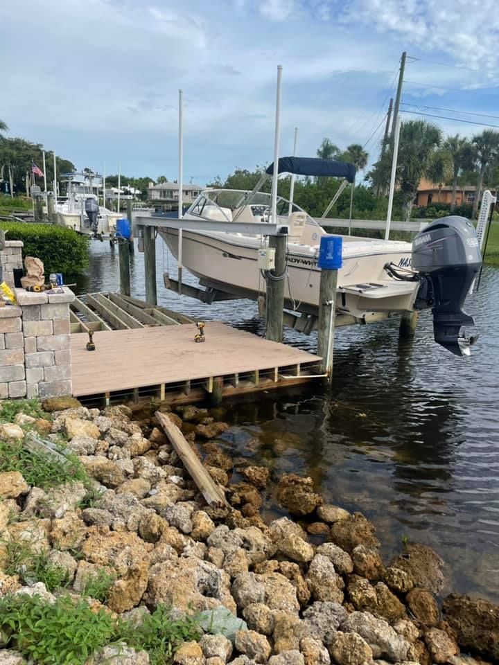 Boat on lift at a dock, wooden platform under construction, rocky shoreline, blue sky.