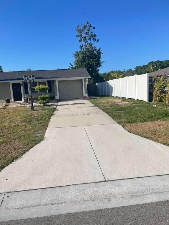 Single-story house with concrete driveway, gray roof, white fence, and blue sky.