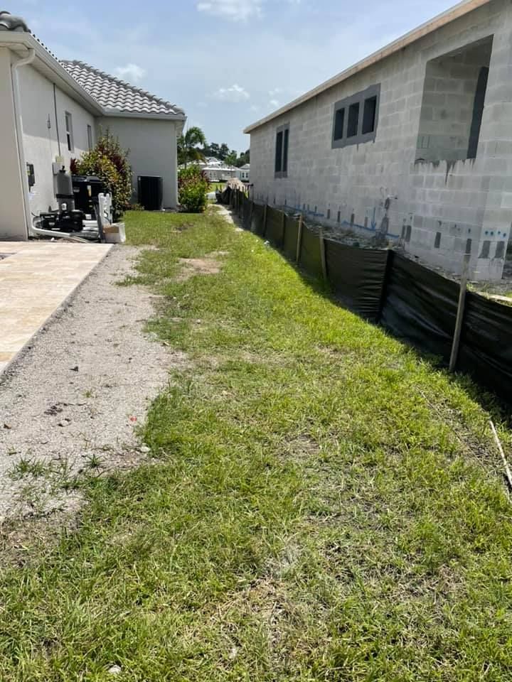 Grassy strip between two buildings, one under construction, with a sandy path on the left.