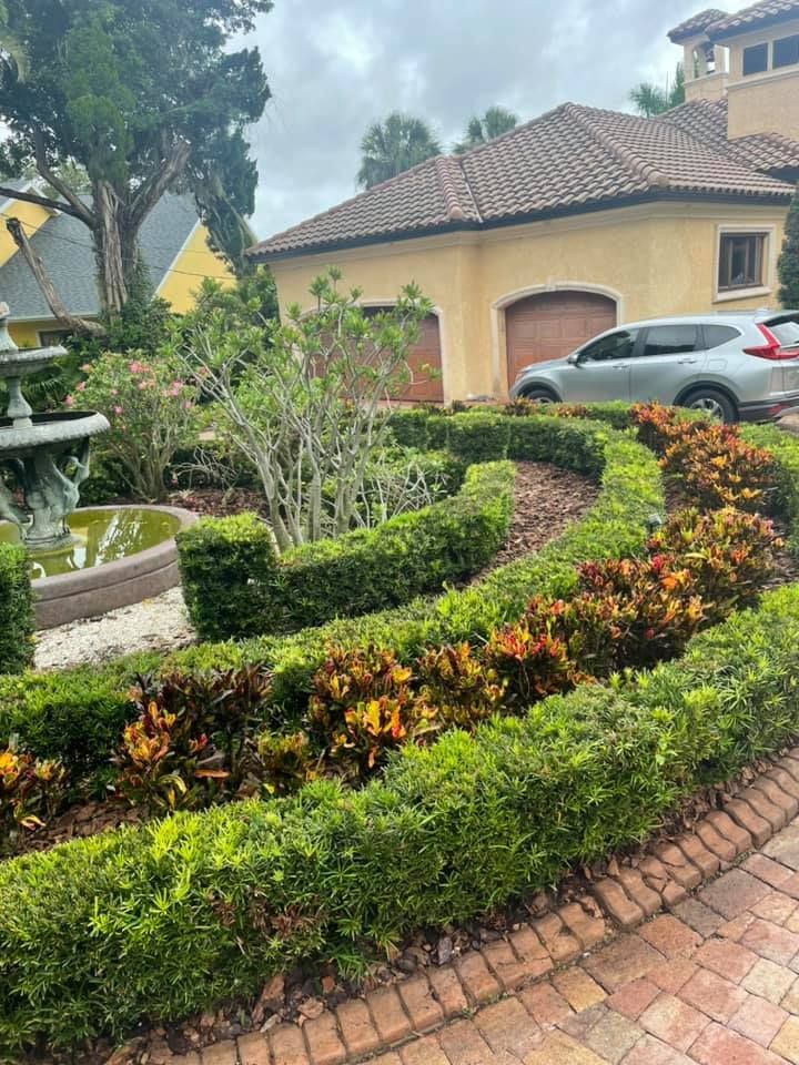 Lush garden beds with manicured hedges and colorful plants curve towards a stucco house with a tiled roof.