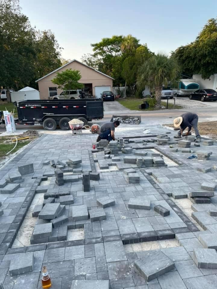 Workers laying gray paving stones in a driveway; a trailer, cars, and houses in the background.