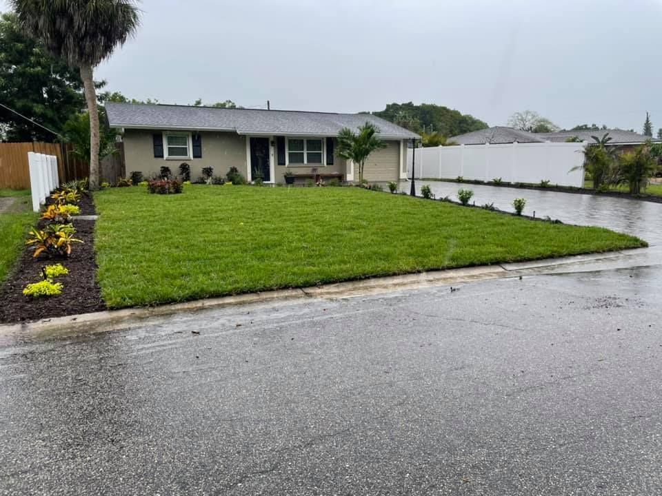 A newly landscaped house with green lawn, flower beds, and a wet, gray street. Overcast sky.