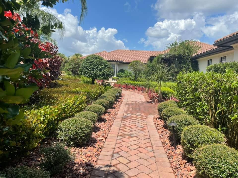 Brick path through a lush garden leads to a large house with a terracotta roof under a partly cloudy sky.