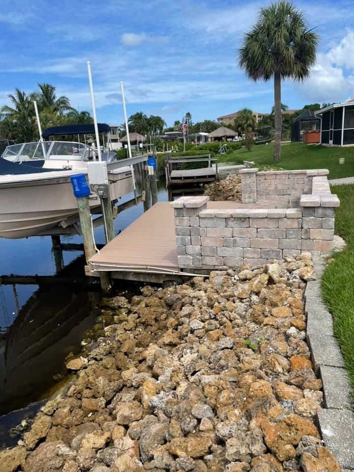 Dock with boat, stacked stone wall, and shoreline rocks under a blue sky with palm tree.