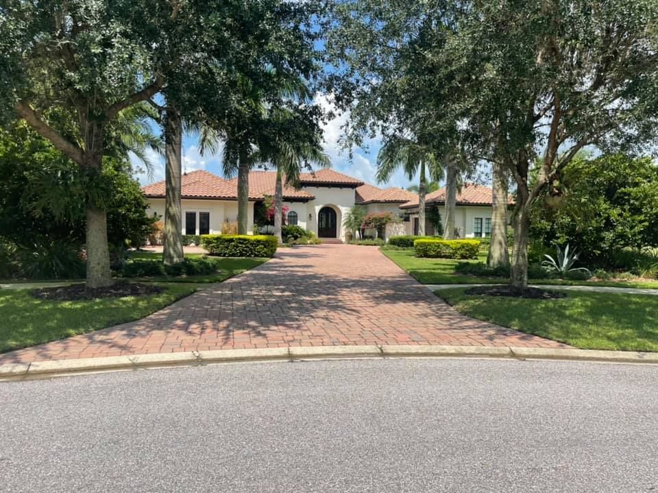 Large brick-paved driveway leading to a beige house with a red tile roof, flanked by trees and green lawns.