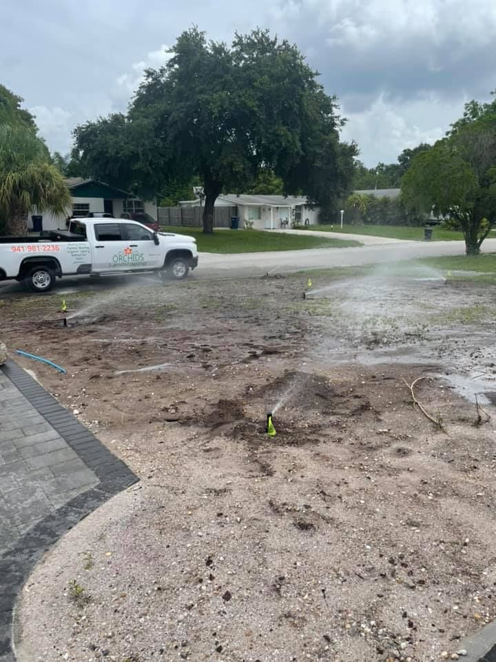Sprinklers watering dirt in a yard; white truck with logo nearby. Houses and a tree in the background.