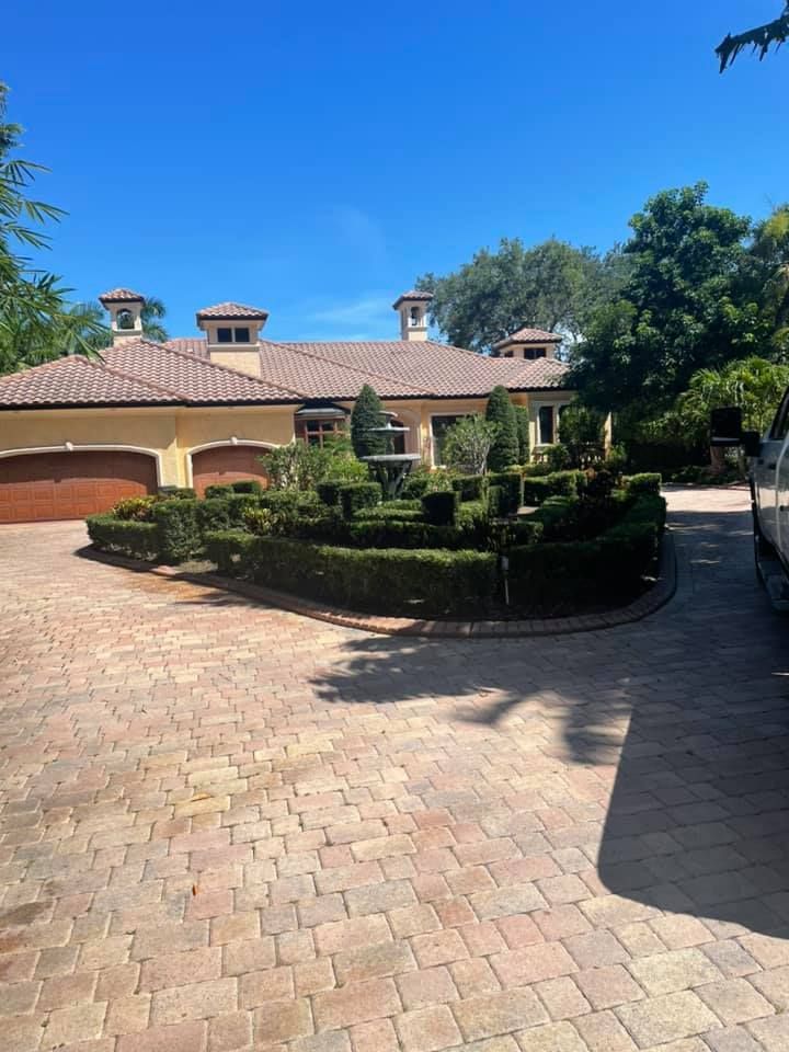 House with a red-tiled roof, yellow walls, and formal hedges surrounding a brick driveway on a sunny day.