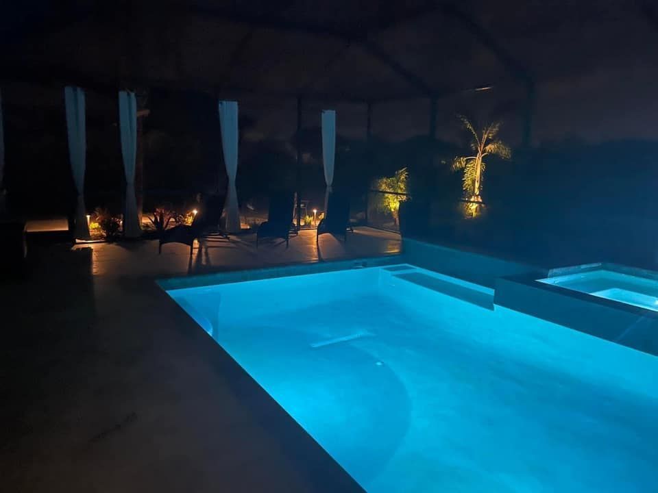 A swimming pool with glowing blue water at night, located on a dark patio with sheer curtains and lounge chairs nearby.