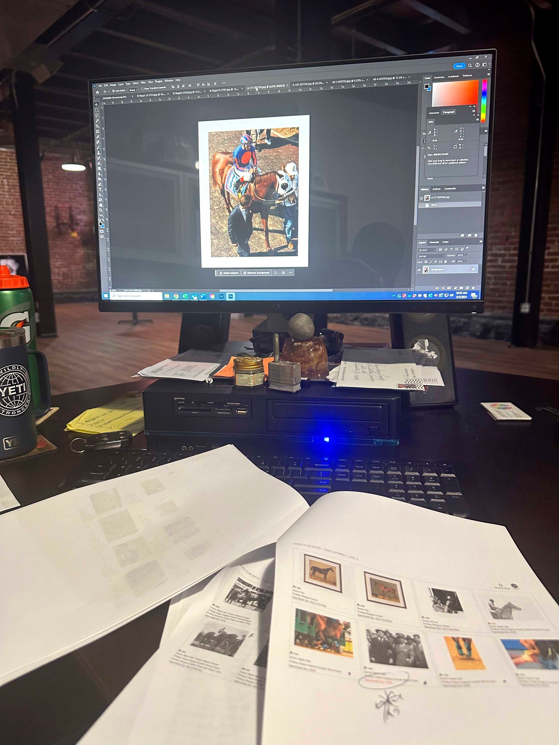 A computer monitor displays an image editing program. Desk with papers, keyboard, and other items in a dimly lit space.