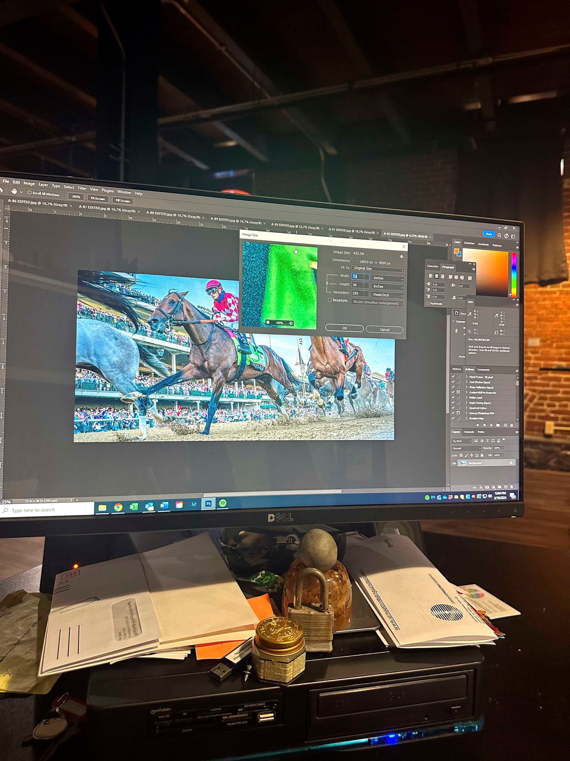 Monitor displaying horse race photo, editing software open. Desk cluttered with papers, objects.