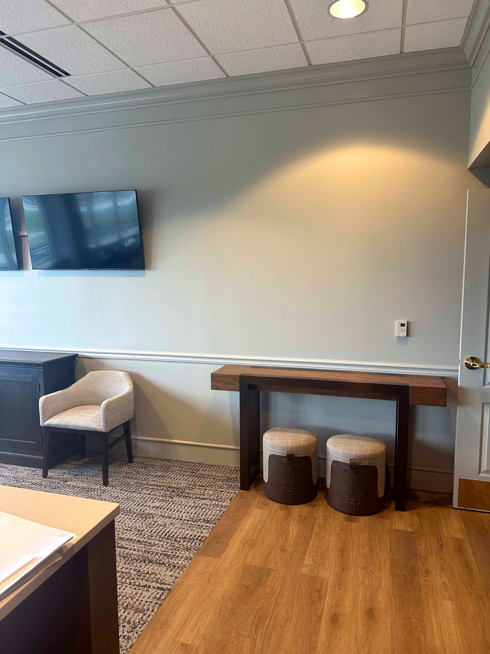 Waiting area with a wooden table, two stools, a chair, and a TV mounted on a light blue wall.