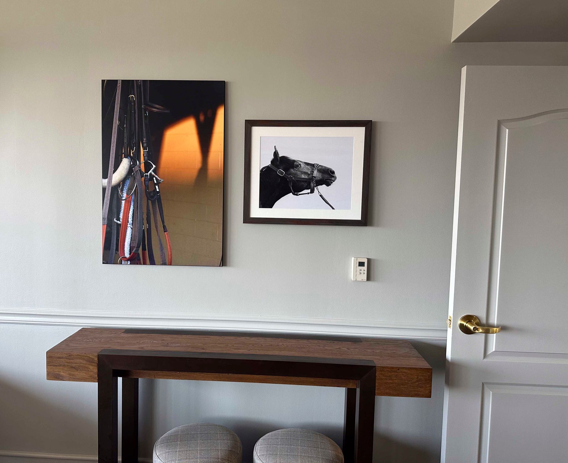 Artwork of horse tack and a horse head are mounted above a wooden console table with two stools. A door is on the right.