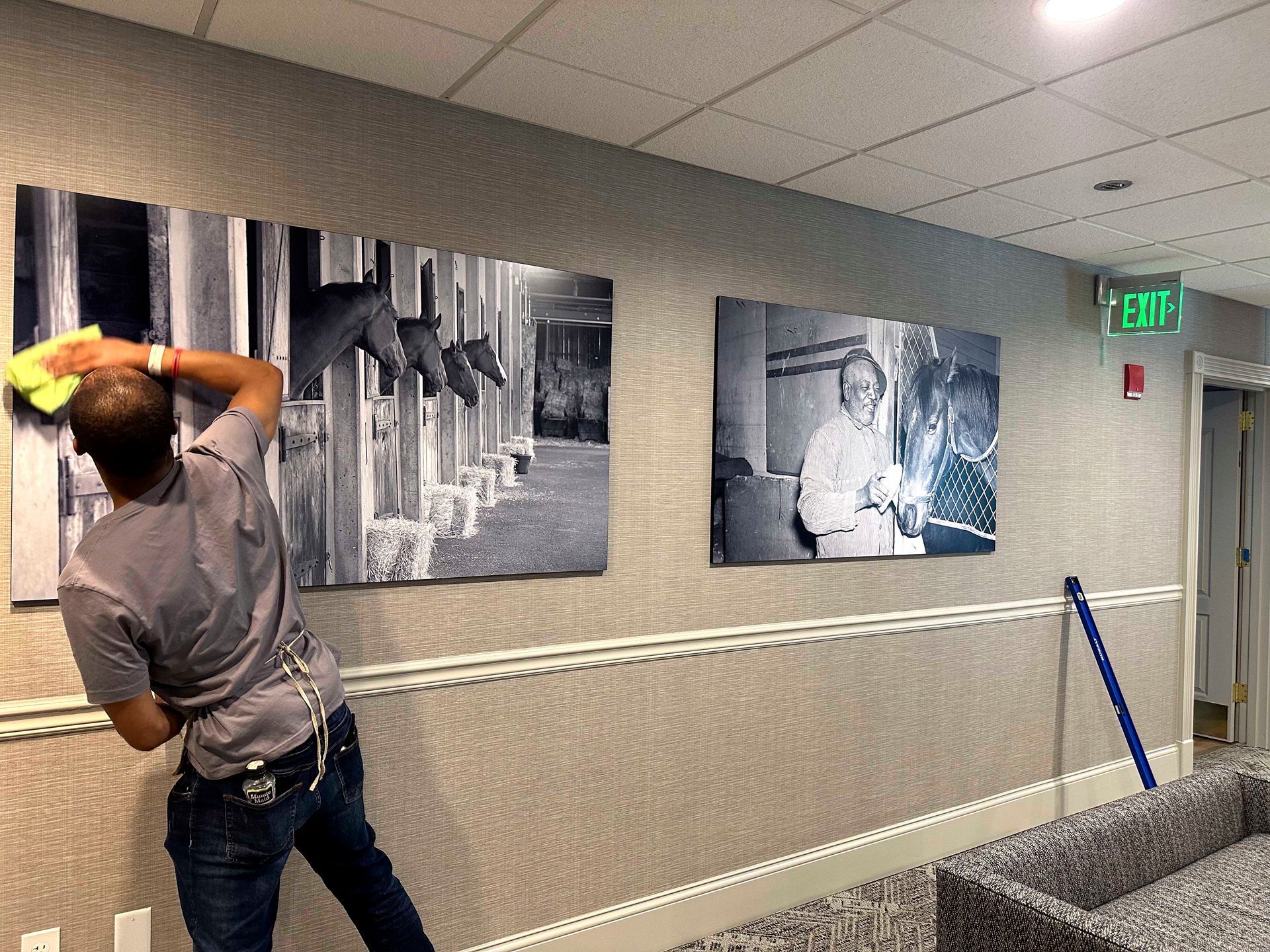 Person cleaning black and white framed photos on a wall in a hallway.