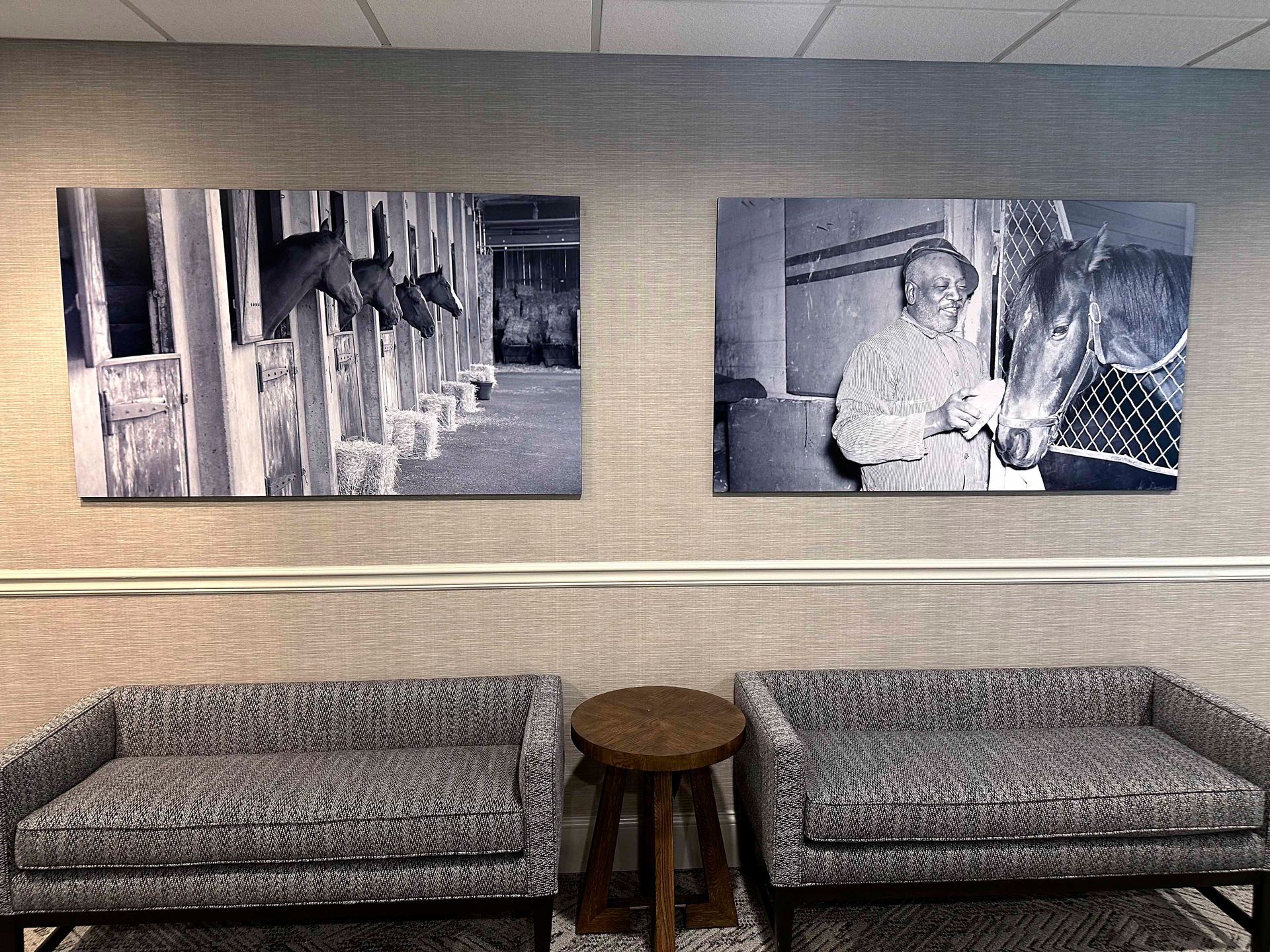 Two black and white photos of horses hanging above two patterned armchairs with a small wooden table between them.