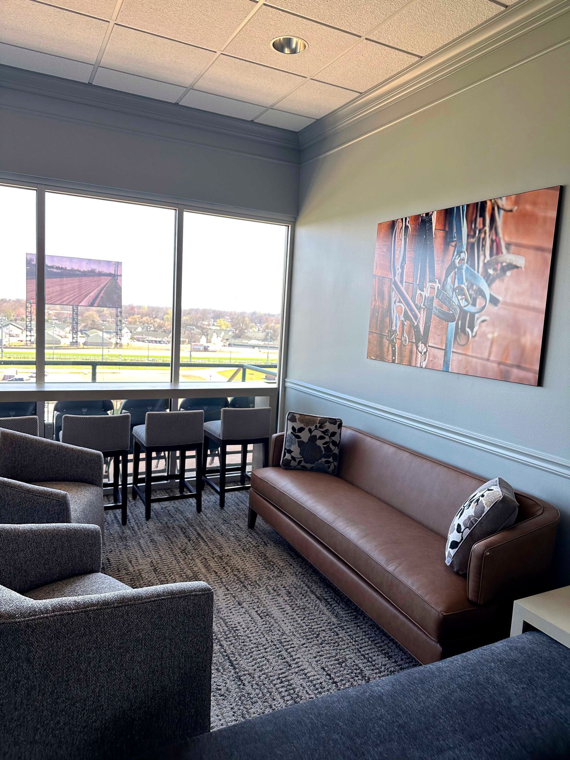 Lounge area with brown leather sofa, artwork, high-top seating by a window. Gray chairs and rug.