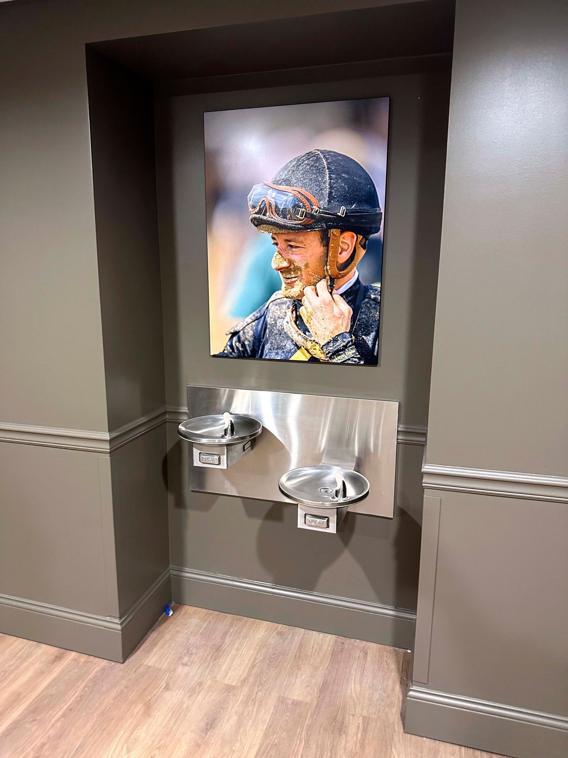 Water fountain below a portrait of a jockey in a building with dark-colored walls and wood floor.