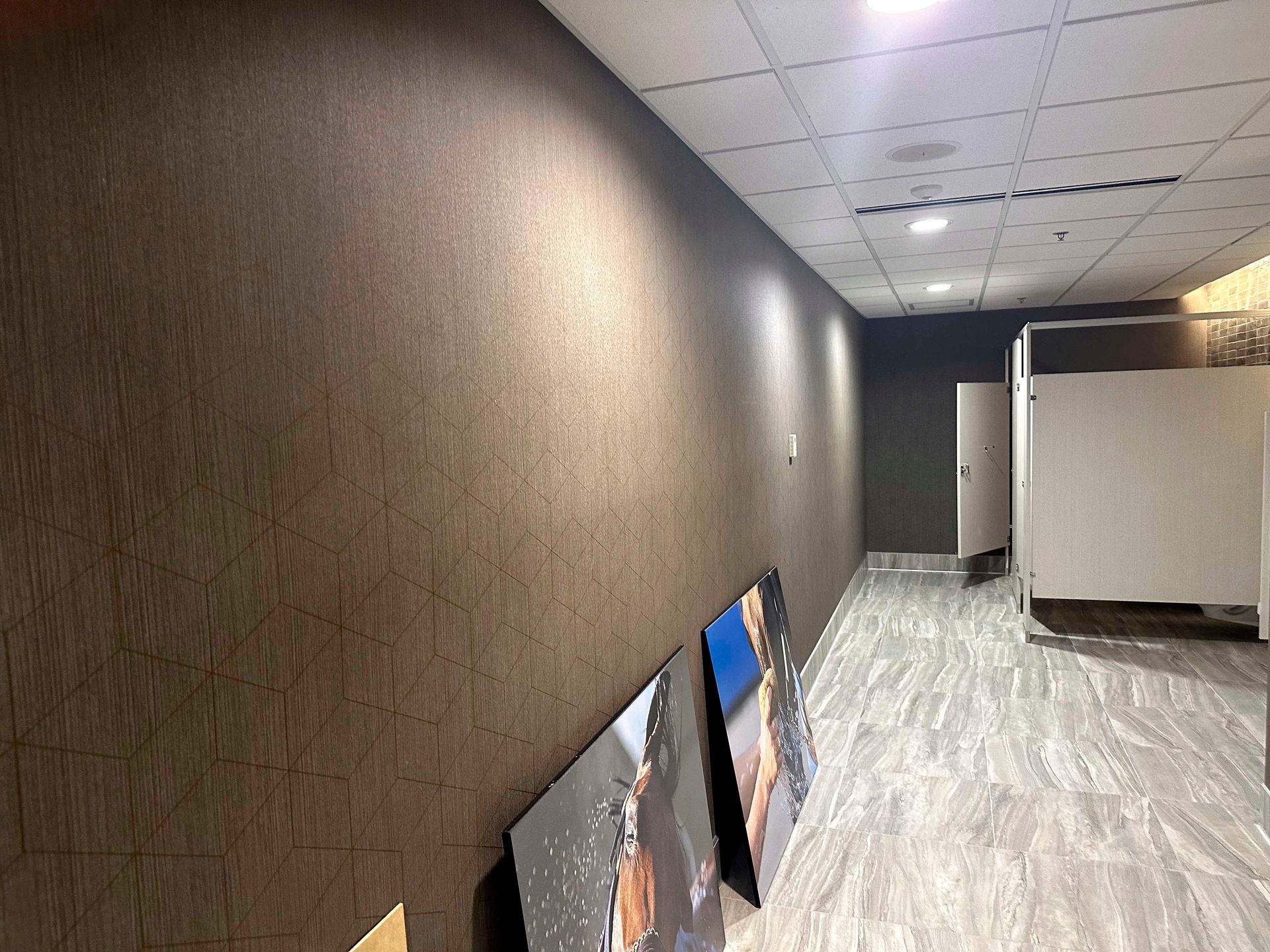 Hallway with textured brown wall, artwork, and bathroom stalls on the right.