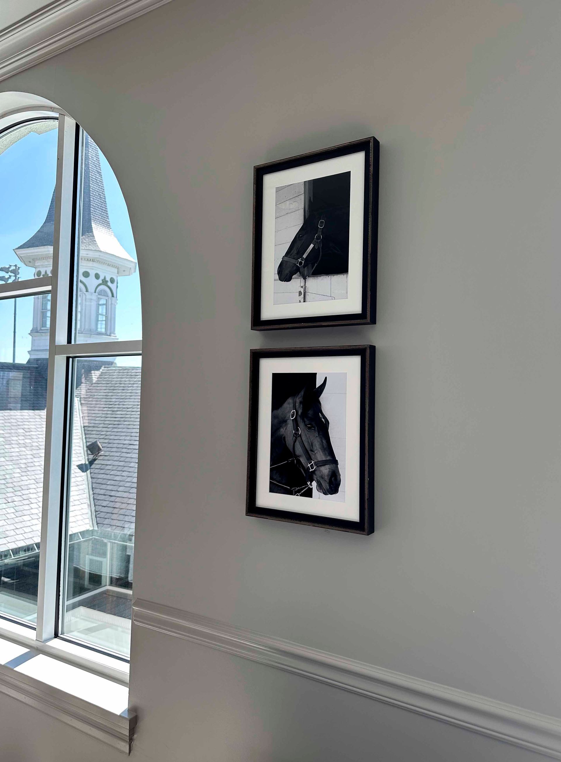 Two framed black and white horse portraits hang on a gray wall next to an arched window.