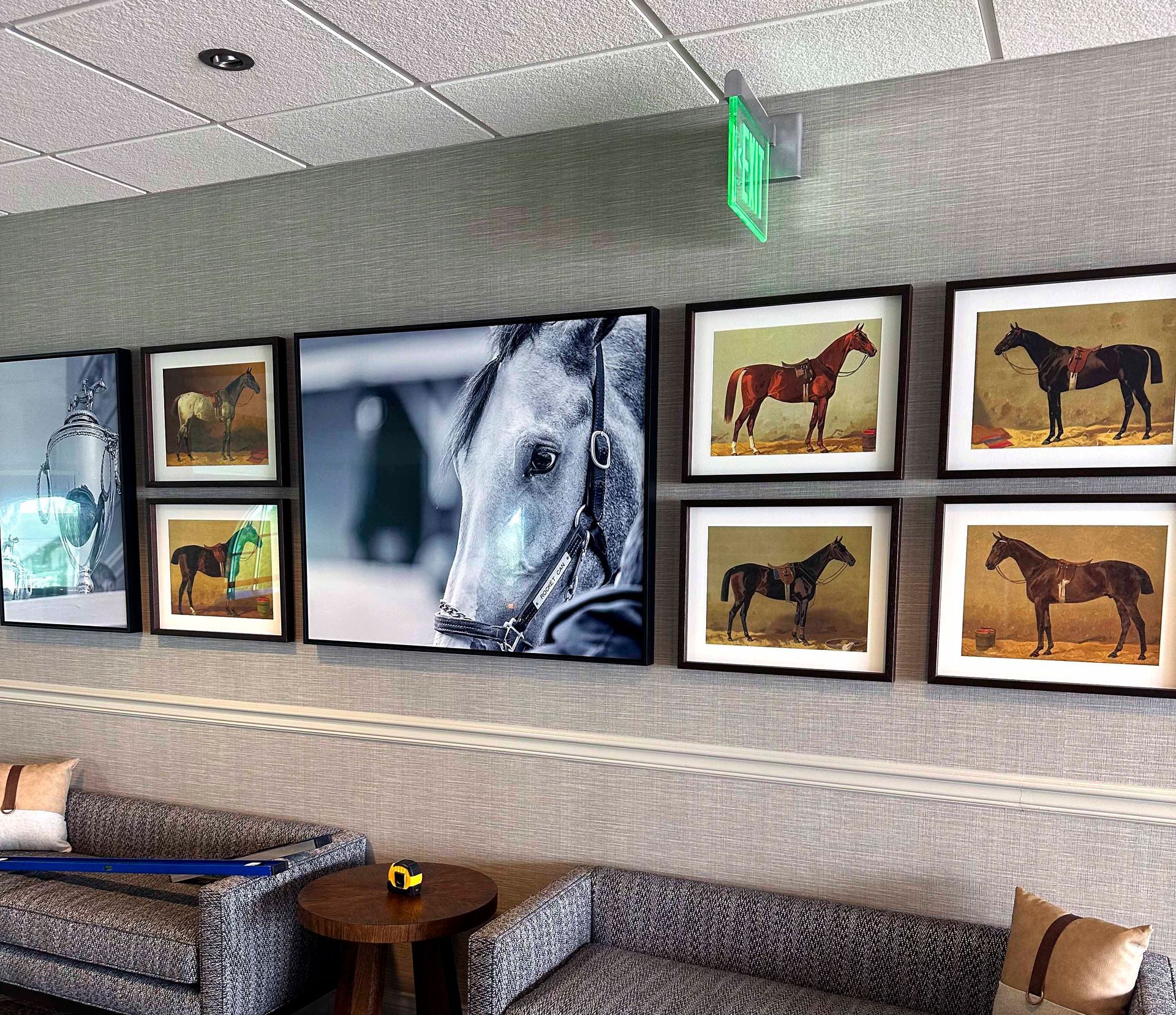 A wall with framed horse art above a sofa. A green exit sign hangs above the art.