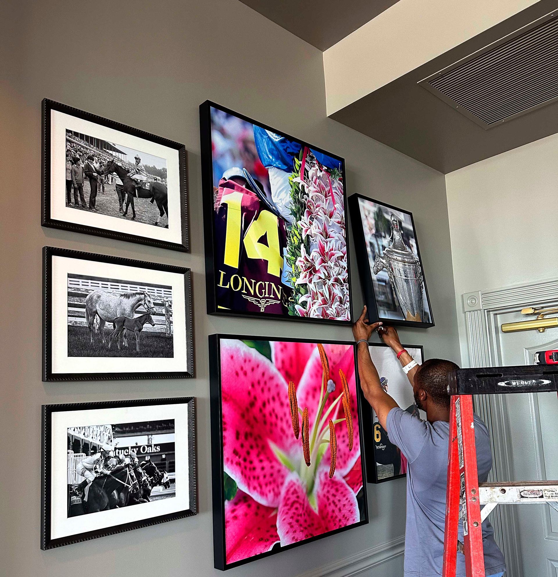 Person hanging framed art on a wall. Includes black-and-white photos and a large, colorful floral print.