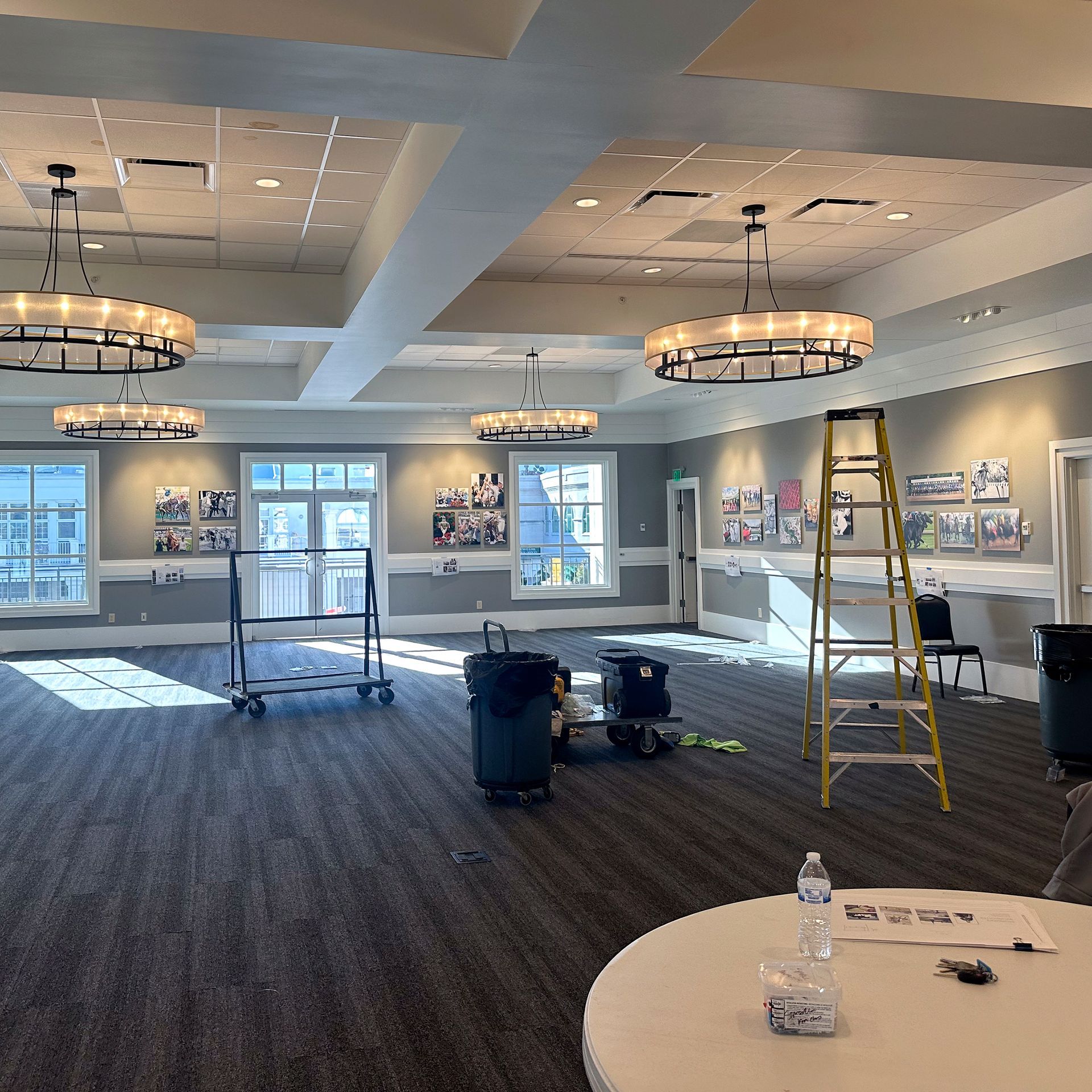 Interior of a large room with round chandeliers, windows, and pictures on the wall. A ladder and cart are in the space.